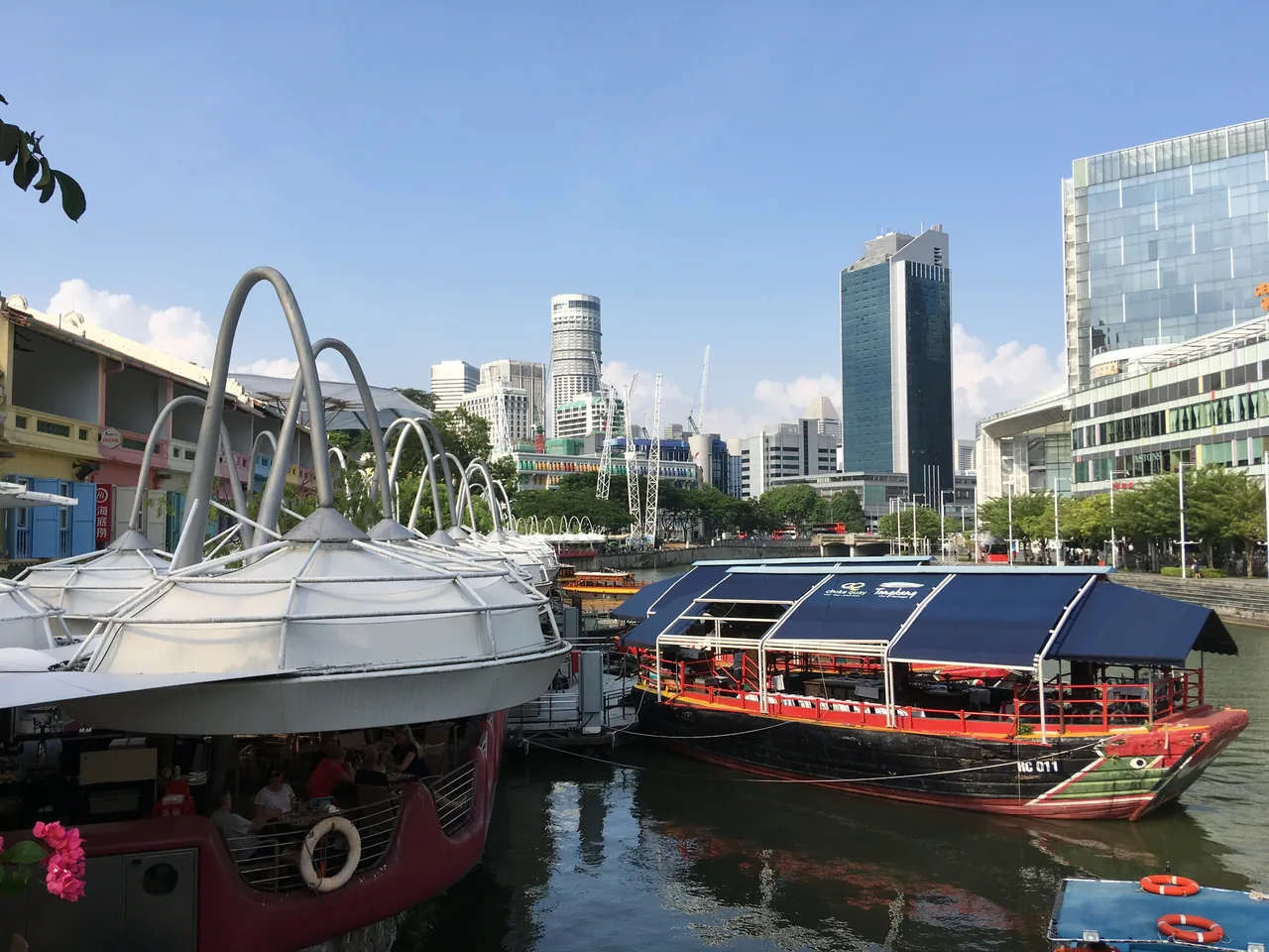 Daytime view of the Clarke Quay riverside in Singapore, with white canopies over the walkway, converted boats moored as floating restaurants, and city towers behind