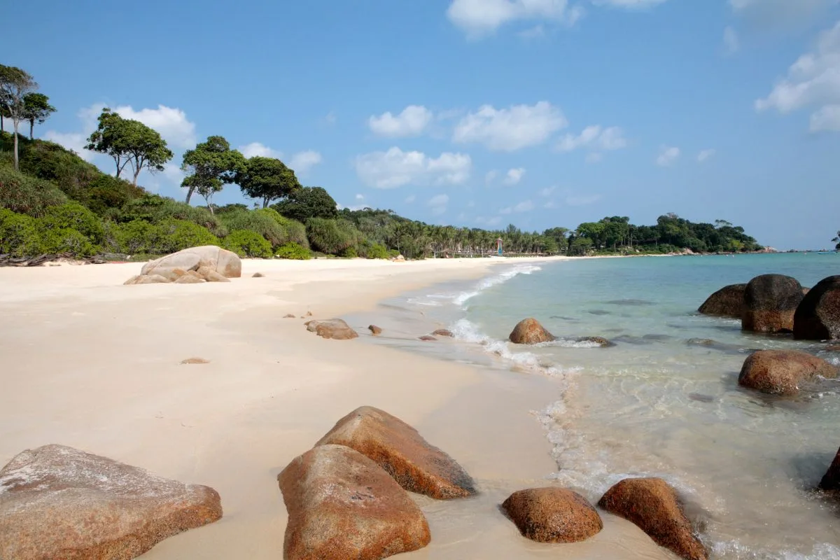 A white sand beach on Bintan with granite boulders in shallow water and trees along the shore