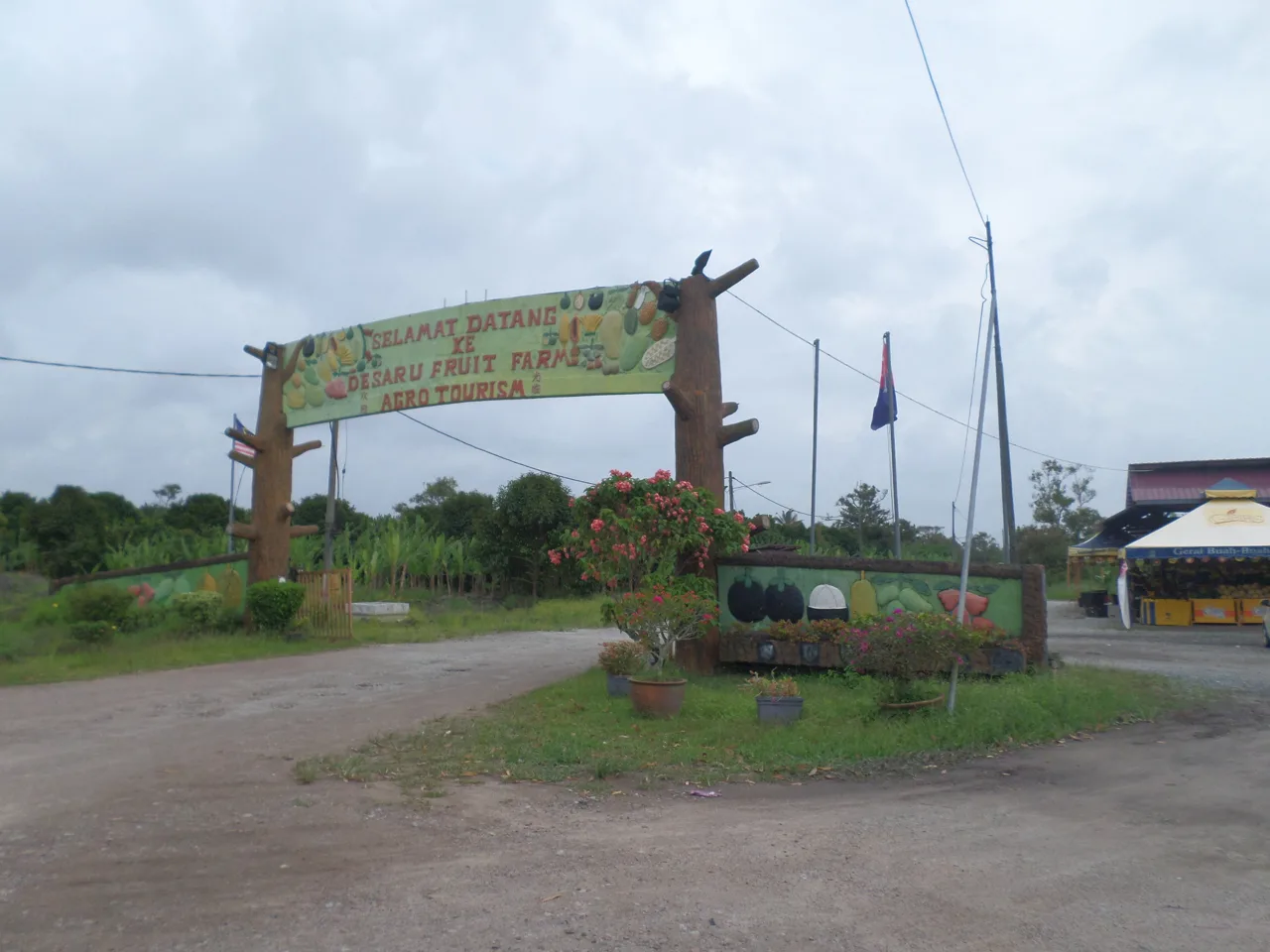 A wooden welcome archway painted with fruit at the entrance to Desaru Fruit Farm, on a gravel road