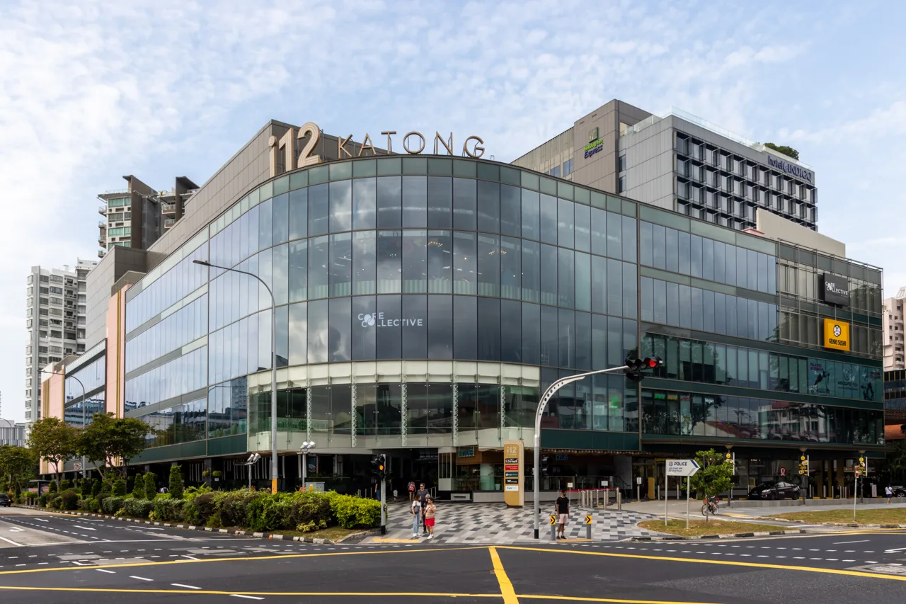 The glass facade of the i12 Katong mall on East Coast Road, with a pedestrian crossing in front and low buildings behind