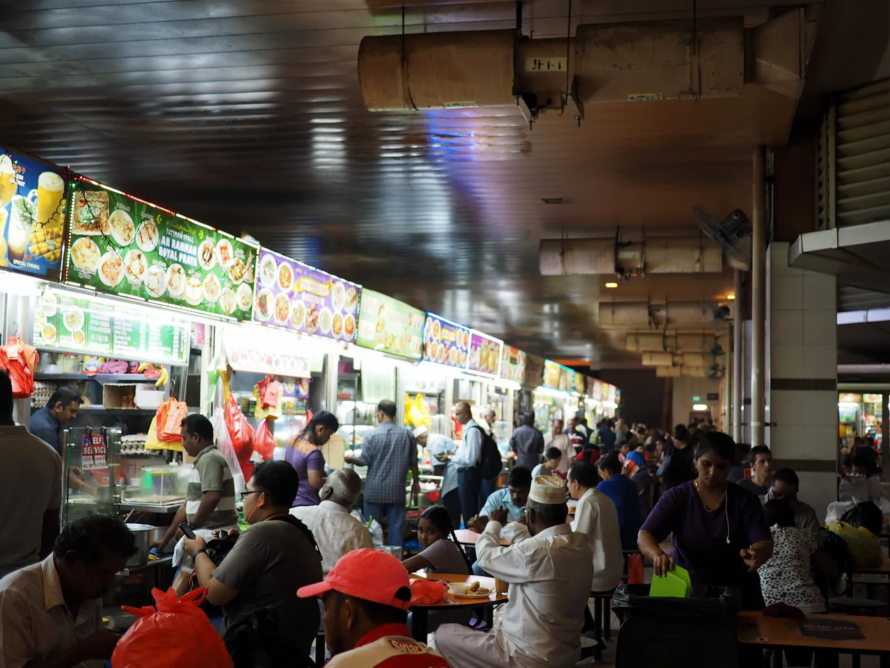 竹脚中心熟食区通道两侧亮着灯的摊位招牌，前方餐桌坐满了人