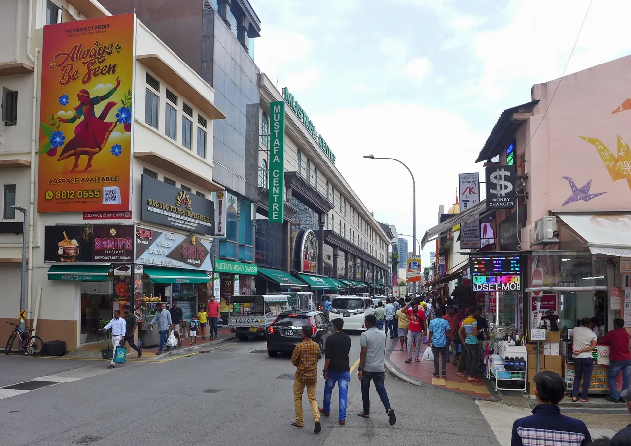 白天挤满行人的街道，尽头的建筑外墙上是绿色的 MUSTAFA CENTRE 直立招牌