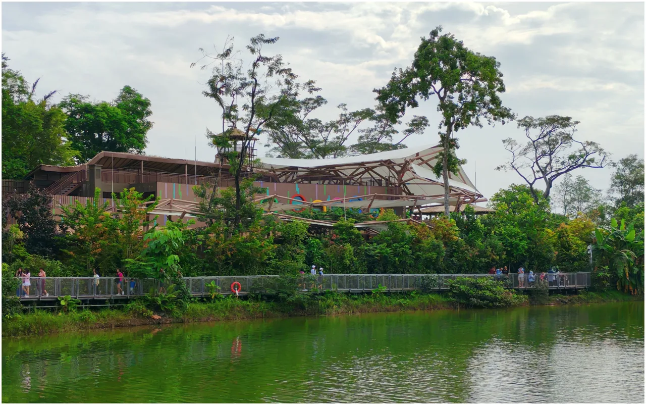 People walking the Mandai Boardwalk deck beside Upper Seletar Reservoir with park buildings behind the trees
