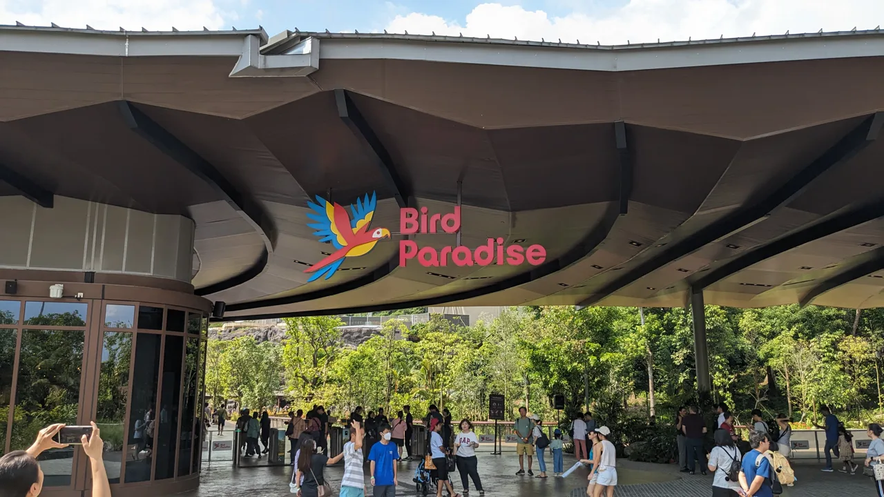 Visitors passing under the sweeping roof of the Bird Paradise entrance beneath its parrot logo