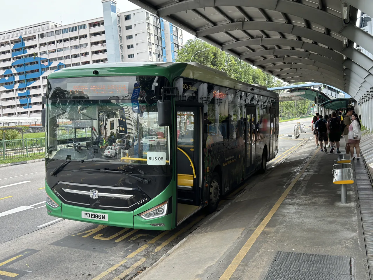 A green Mandai Khatib Shuttle bus at the stop with passengers boarding under a covered walkway