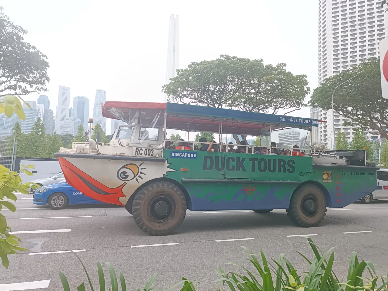 A Singapore DUCKtours amphibious vehicle painted with a duck bill and eye, carrying passengers along a road with the city skyline behind