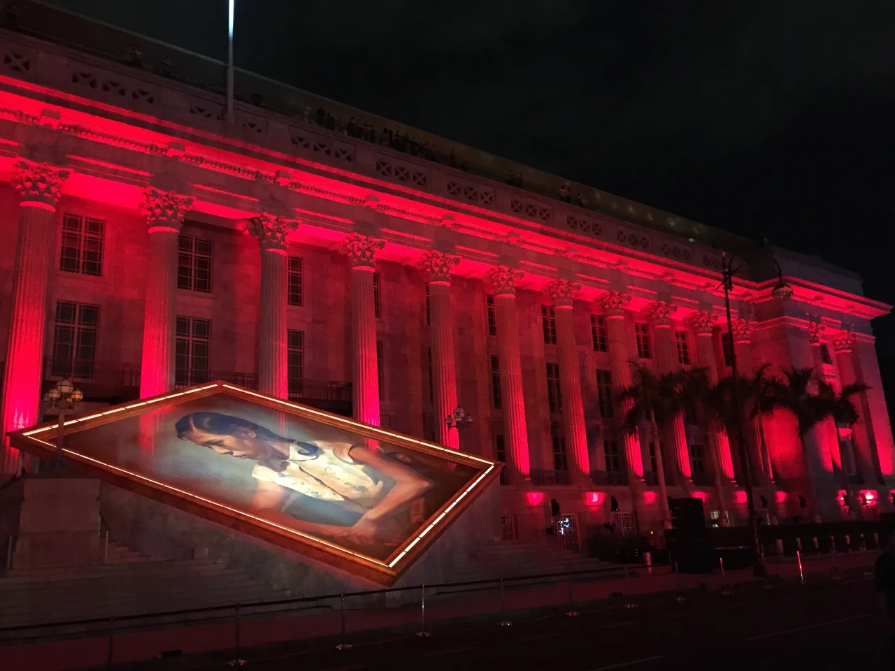 The columned facade of the National Gallery Singapore lit red at night with a large painting projected in front of the steps