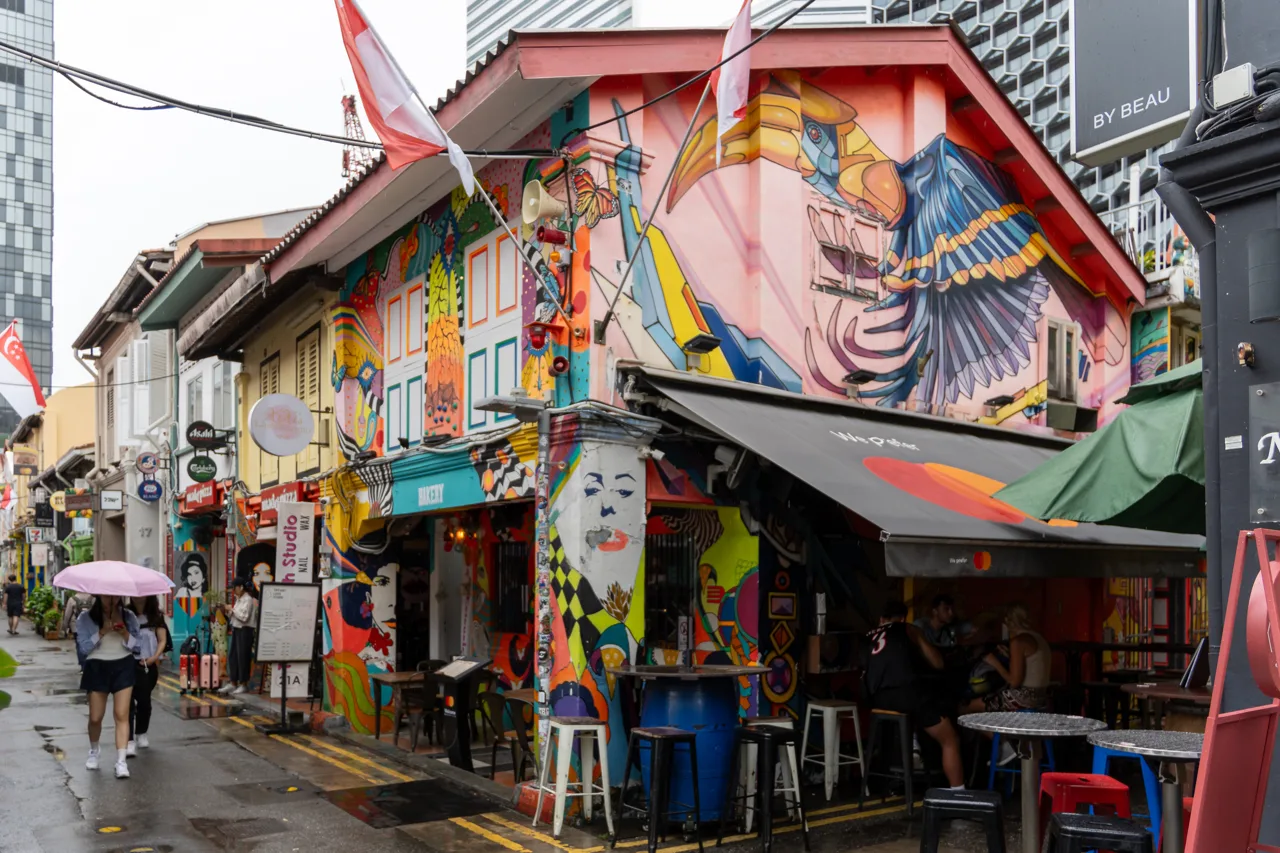 A narrow Singapore lane lined with shophouses covered in murals, with people walking past bar stools set out on the wet street