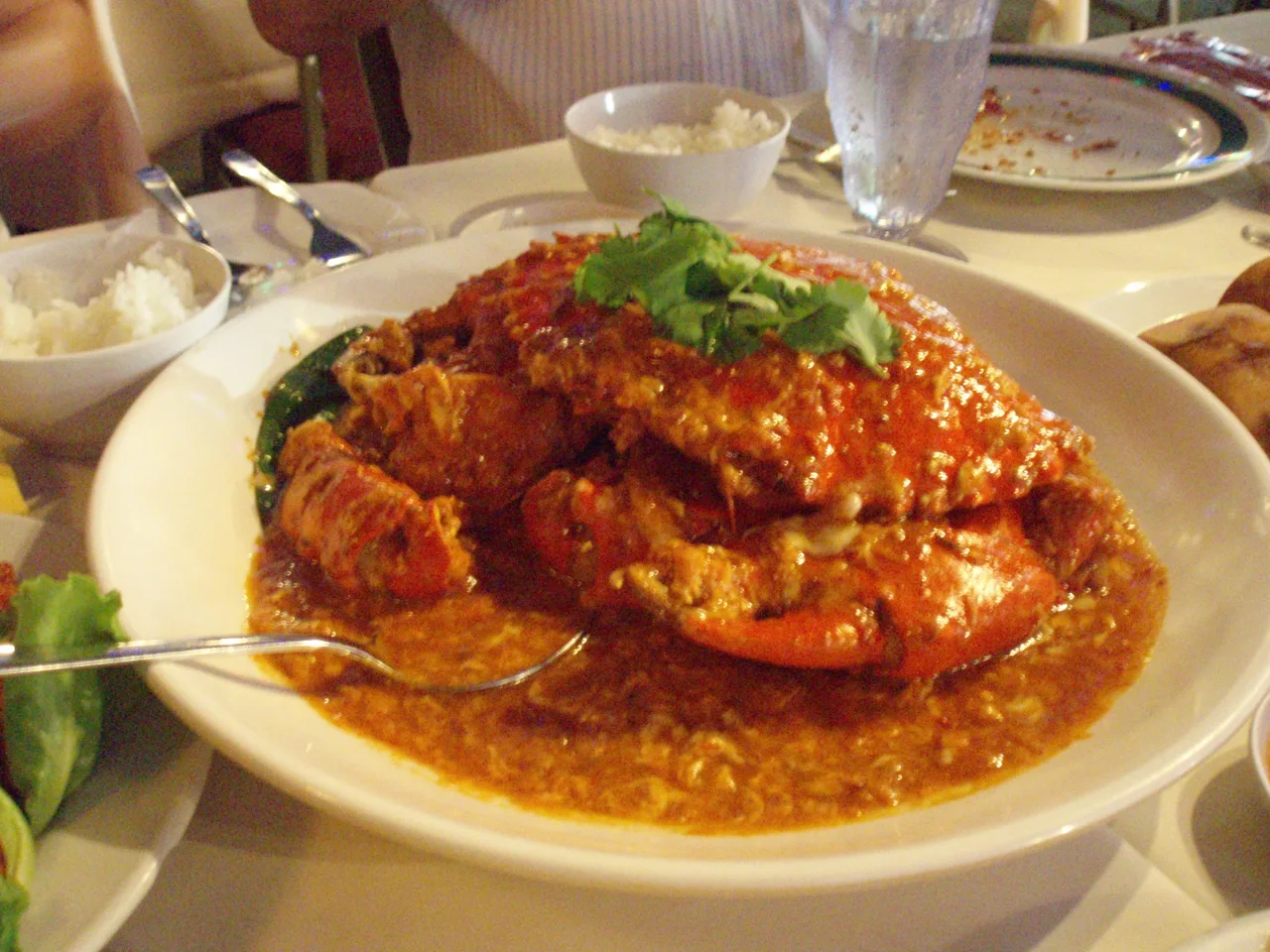 A whole chilli crab served in thick red sauce on a large white plate, with two bowls of rice beside it