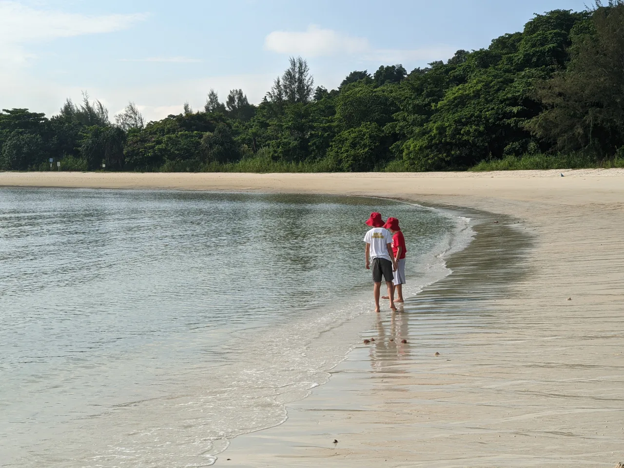Two people walking along the curving white sand of Lazarus Island beach with dense green forest behind them