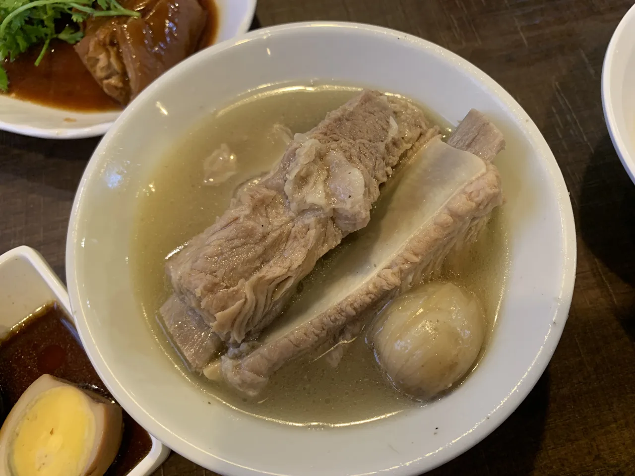 A white bowl of clear broth holding two pork ribs and a whole garlic clove, with a dark braised dish and half a boiled egg on plates alongside