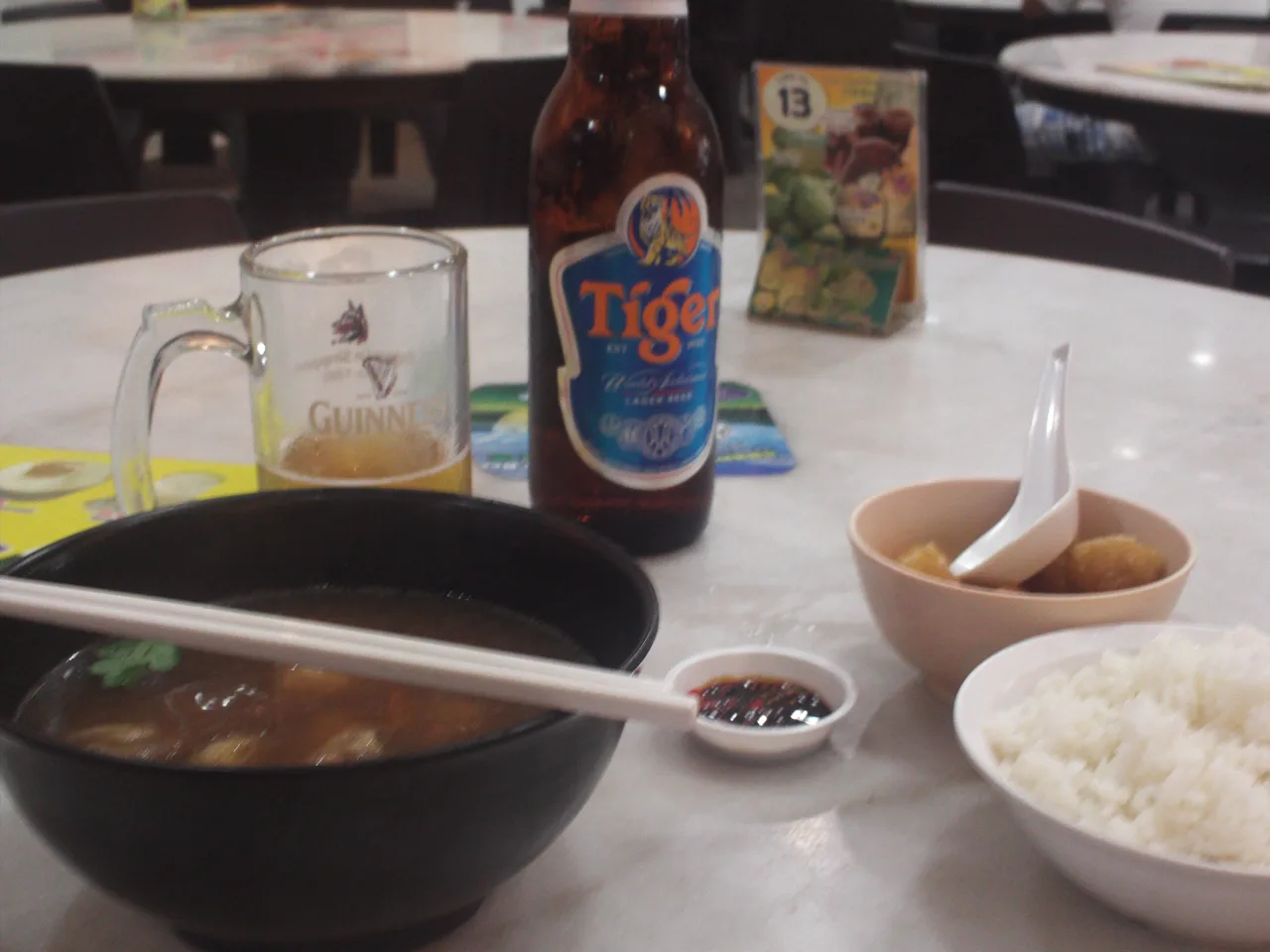 A black bowl of darker broth beside a bowl of cut pieces of you tiao, a bowl of white rice, a saucer of dark soy sauce, a bottle of Tiger beer and a glass.
