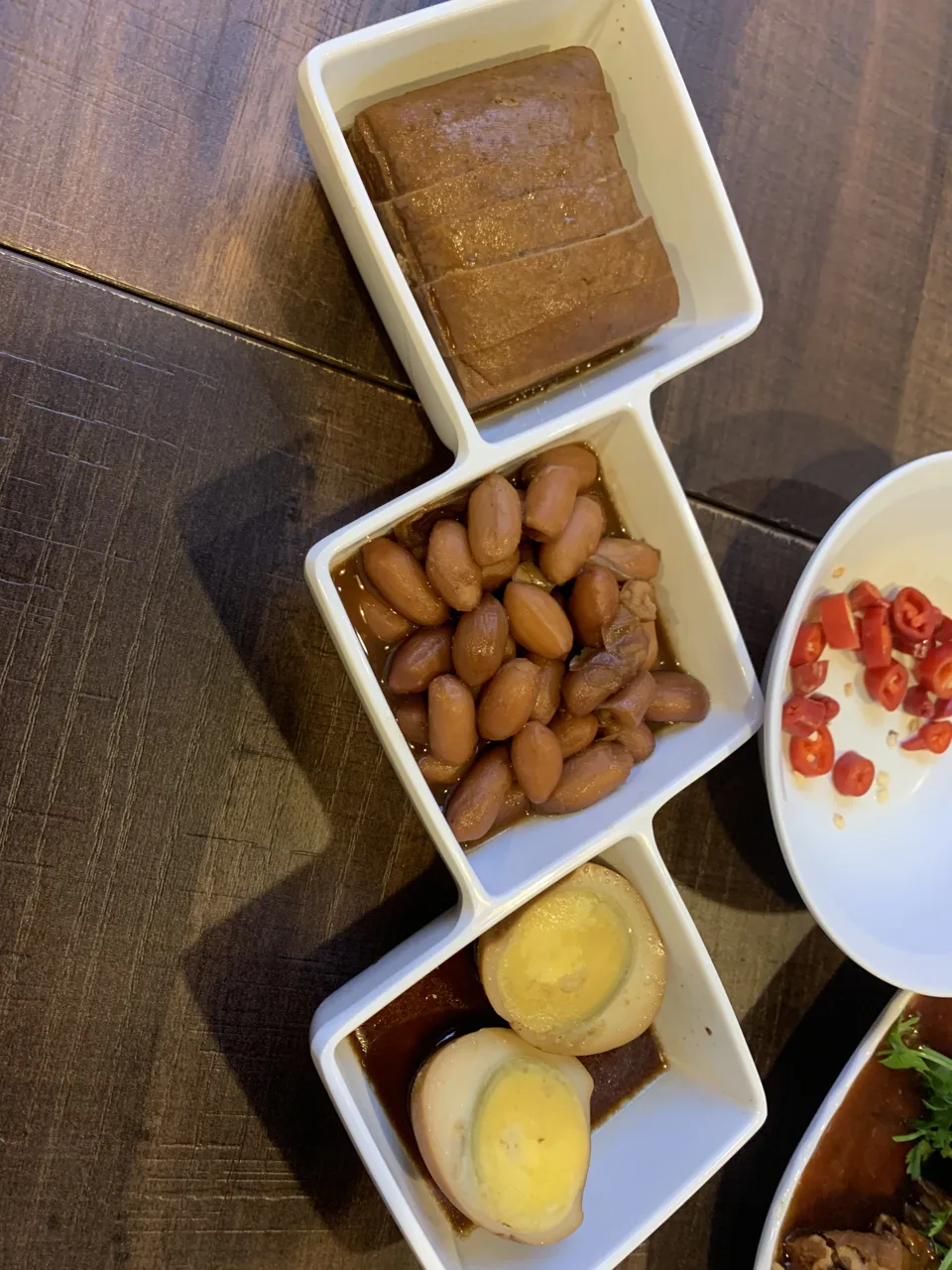 A white dish with three compartments holding braised beancurd, peanuts and halved boiled eggs, next to a side plate of sliced red chilli.