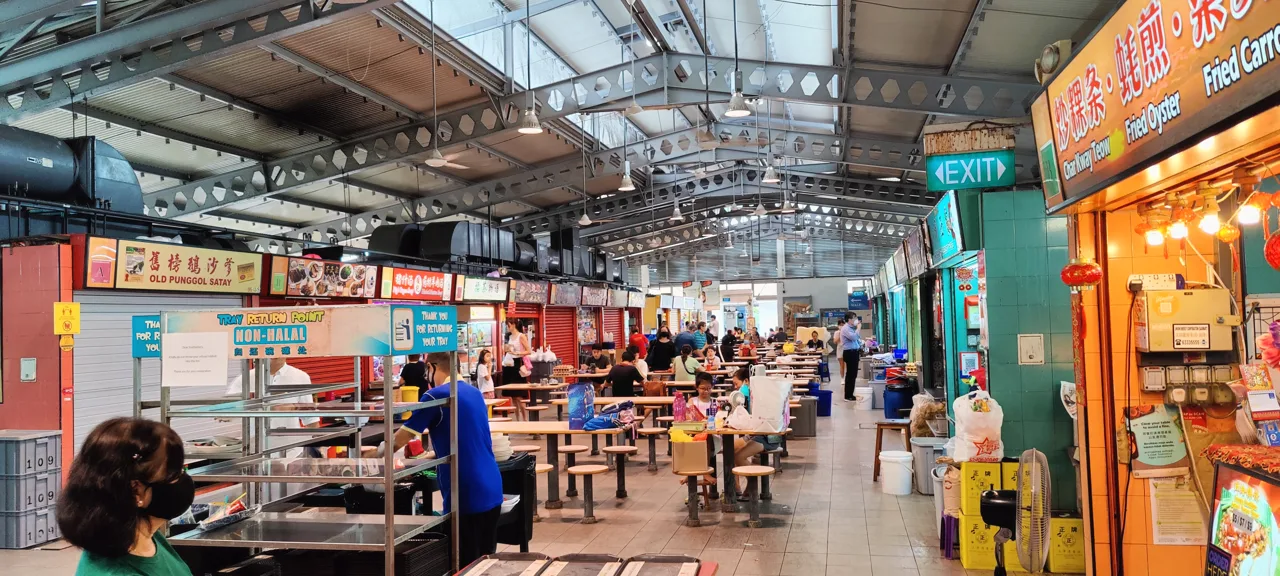 The inside of Alexandra Village Food Centre, with stall signboards, communal tables whose round stools are fixed to the frame, and a sign reading Tray Return Point NON-HALAL.