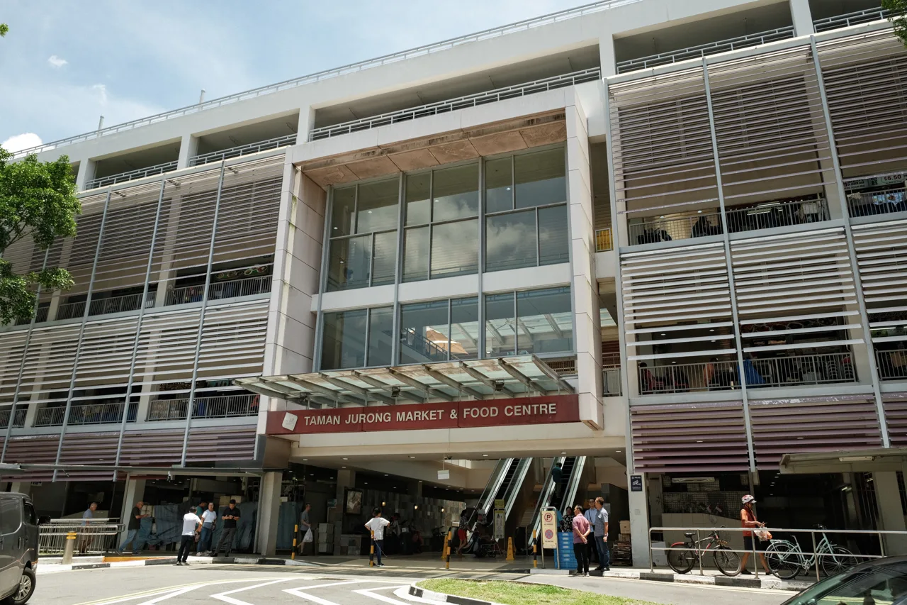 The outside of Taman Jurong Market and Food Centre, showing the building signboard, escalators, parked bicycles and people coming and going.