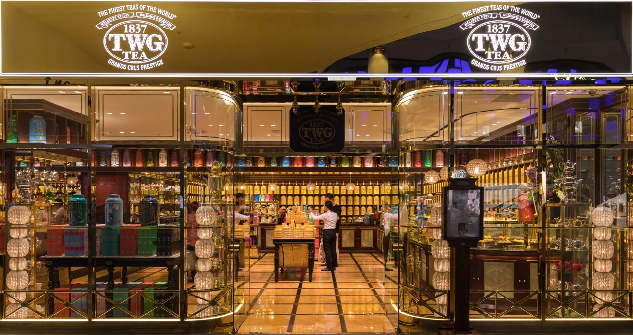 The lit frontage of a TWG Tea shop, with 1837 TWG TEA on the sign above the entrance and shelves of tea tins behind the glass