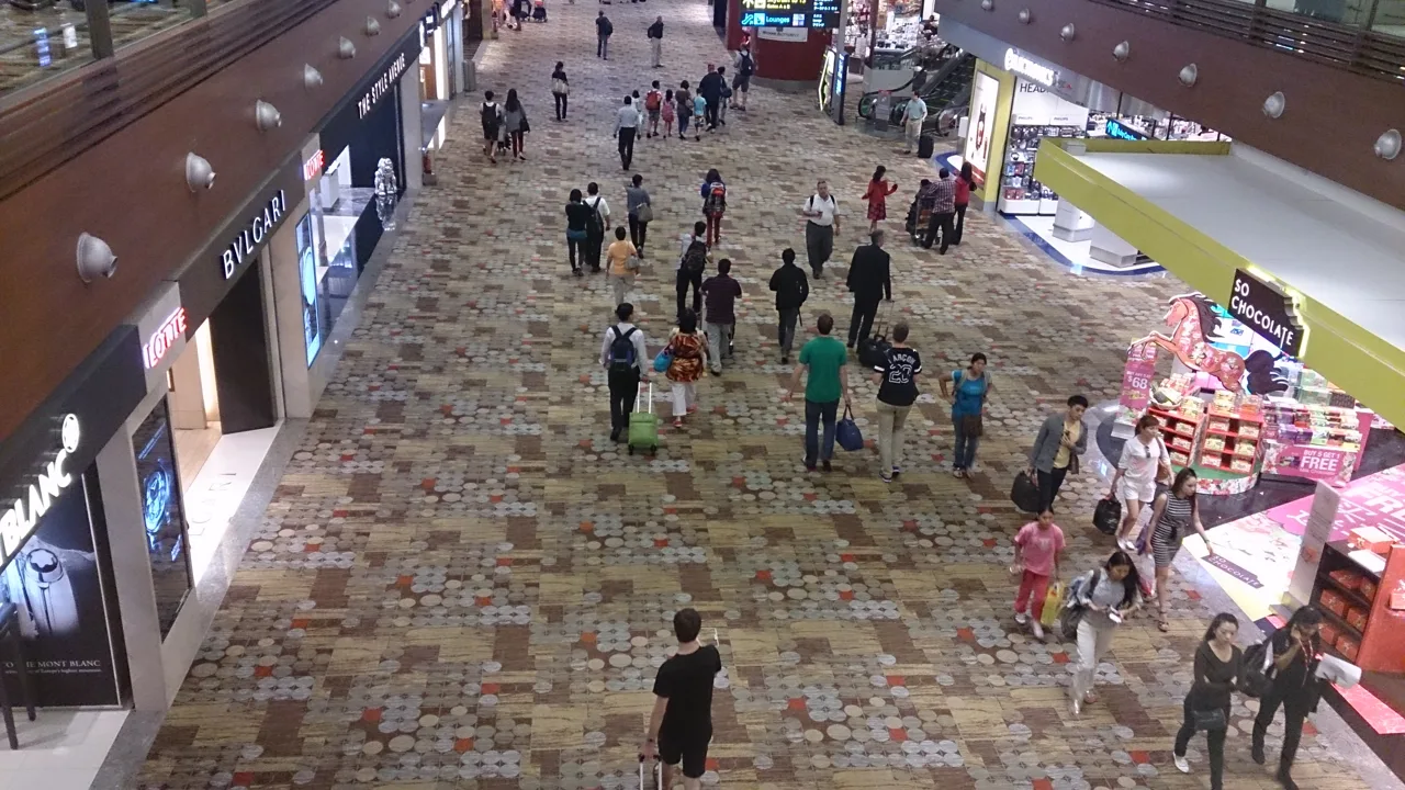 Travellers walking through the Terminal 1 transit hall at Changi Airport, past boutiques on one side and a confectionery stand on the other