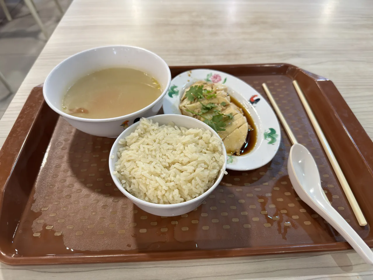 A tray holding a bowl of soup, a separate bowl of rice and a plate of poached chicken with dark soy and coriander, with chopsticks and a spoon