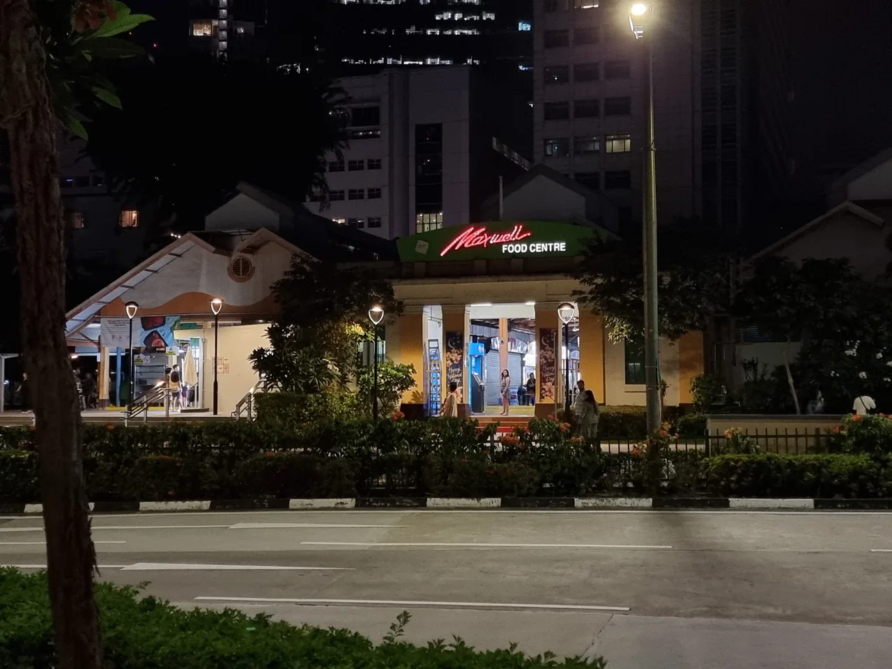 Maxwell Food Centre at night, seen from across the road, with a red neon sign on the building and planting and a carriageway in front of it