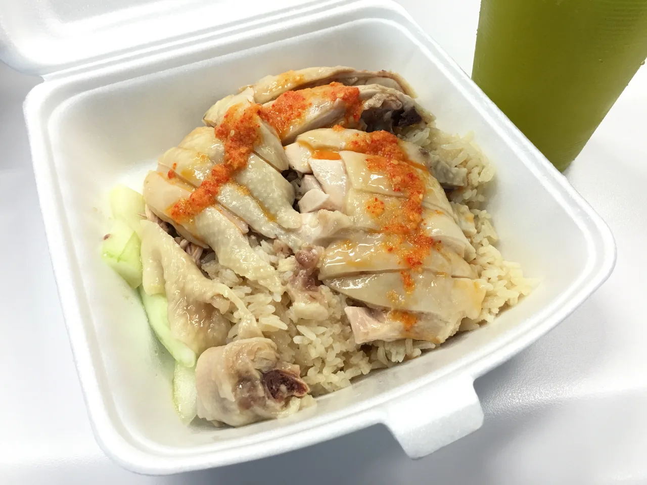 Tian Tian chicken rice in a polystyrene takeaway box, with poached chicken over rice, chilli sauce and a few slices of cucumber, and a green drink cup standing beside it