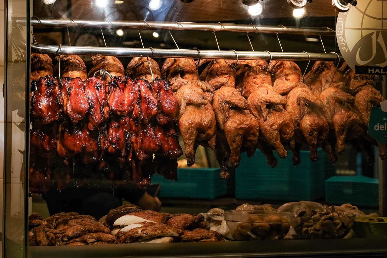 Roast meats hanging in a glass display case, red char siew on the left and roast birds on the right, with a halal sign at the top right of the case