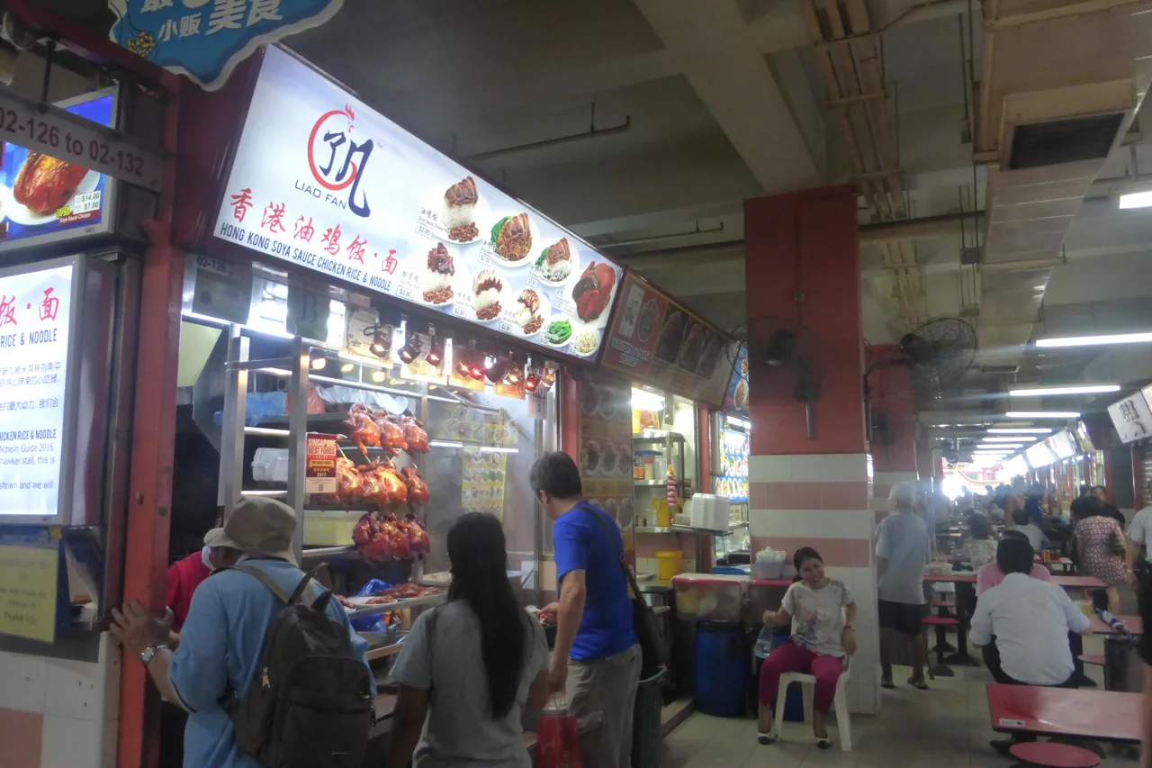 The Liao Fan stall front in Chinatown Complex, with its signboard above the counter, soy chickens hanging in the window, a few customers at the counter and hawker seating behind them