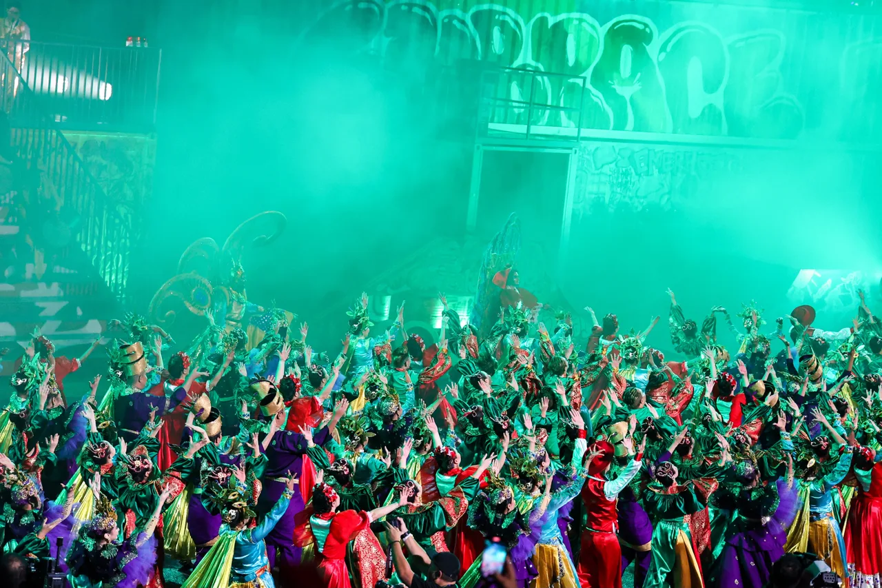 Dozens of Chingay performers at night in green and red costumes with their arms raised, lit by green light and smoke