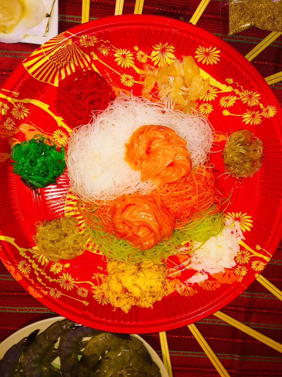 A red platter of yusheng with raw salmon and shredded vegetables arranged in blocks of colour, chopsticks resting around the rim, not yet tossed