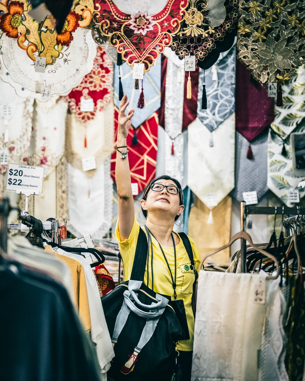 A stall of embroidered table runners at the Geylang Serai bazaar, with a shopper in yellow reaching up towards them