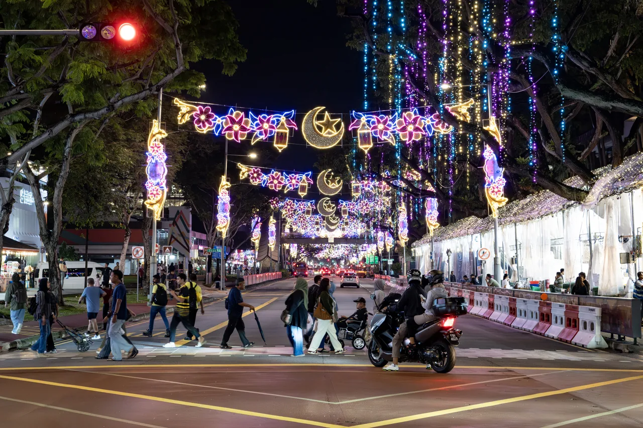 Lampu hias bulan sabit dan bintang di atas jalan Geylang Serai pada malam hari dengan tenda bazar di sisi jalan