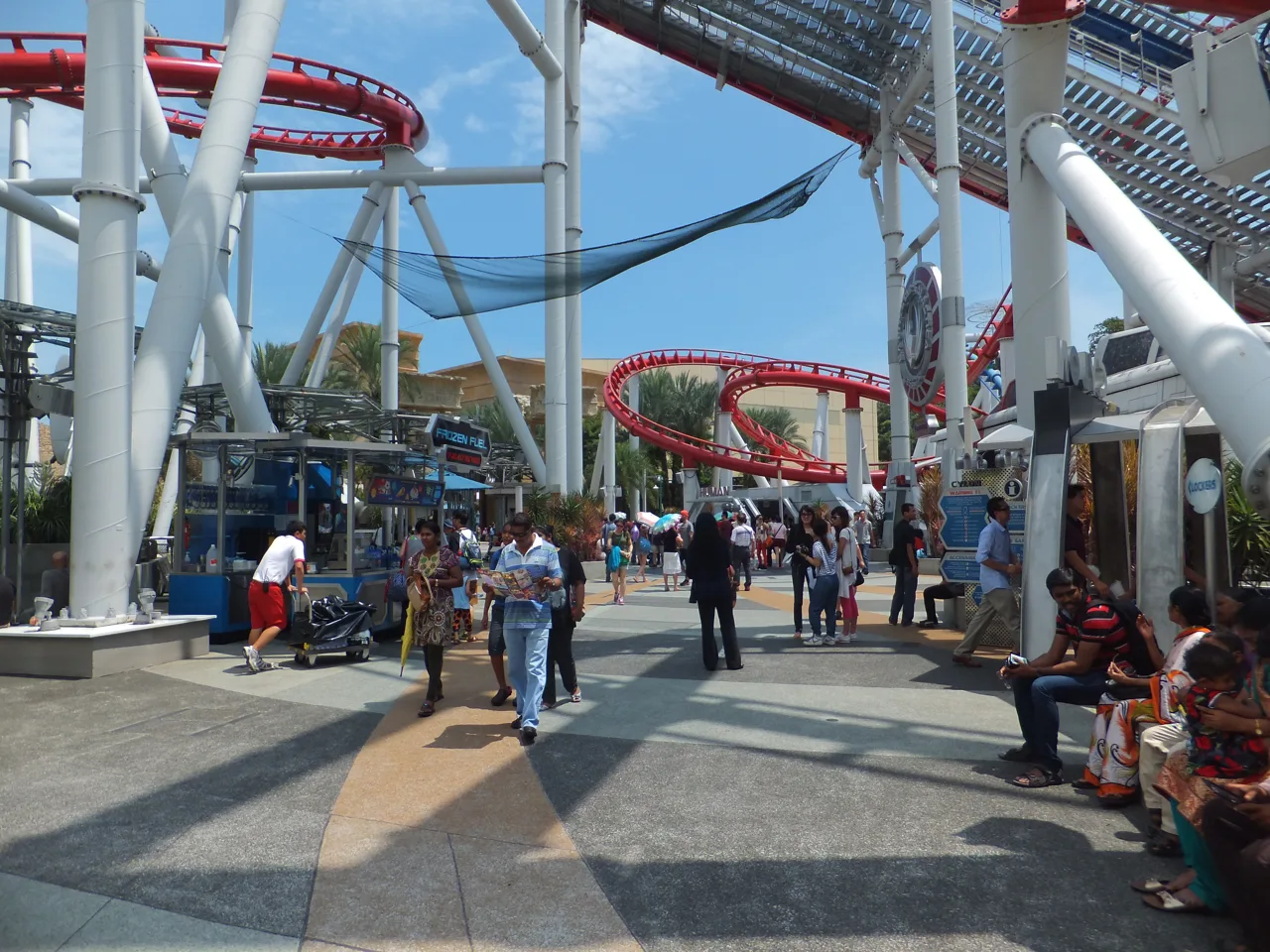 Pengunjung berjalan dan duduk di bawah rel roller coaster merah-putih di Universal Studios Singapore
