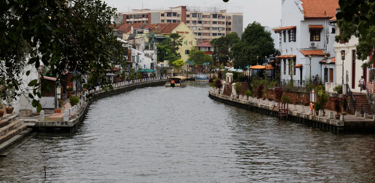A cruise boat heading up the Melaka River between riverside cafes and low heritage buildings on both banks