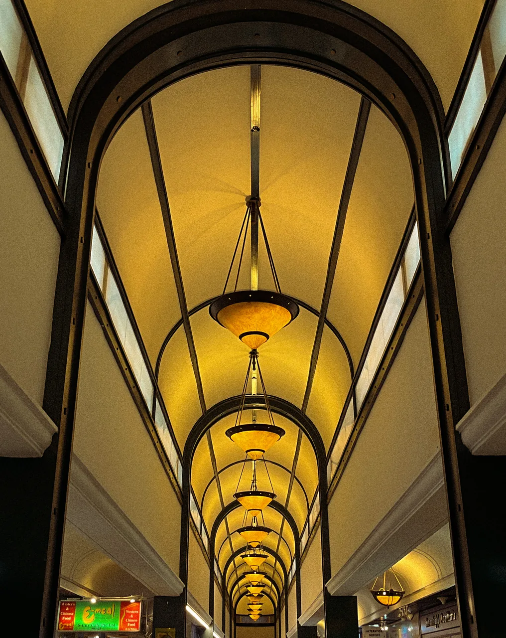 Looking up inside The Arcade in Singapore: an arched ceiling with a row of hanging lamps above the money changer shopfronts below