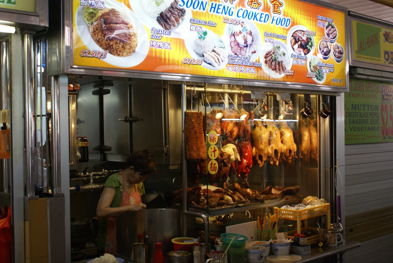A hawker stall front with a lit menu board, roast meats hanging in the display case and a cook working behind the counter