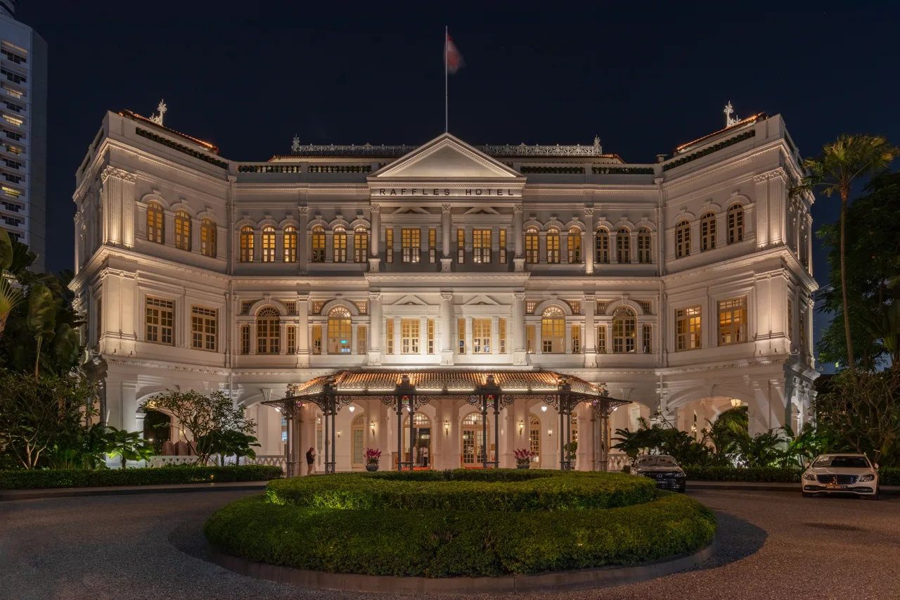 The floodlit front of Raffles Singapore at night, with the covered carriage porch at the centre and a circular lawn in the driveway