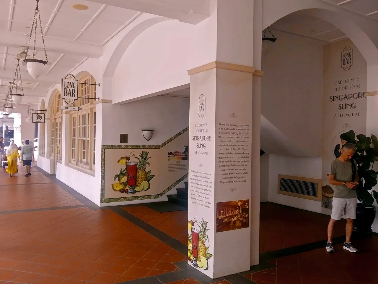 An arcade walkway with hanging lamps along the ceiling, a bracket sign reading LONG BAR THIS WAY, a staircase up to the next level, and a pillar panel painted with a Singapore Sling.