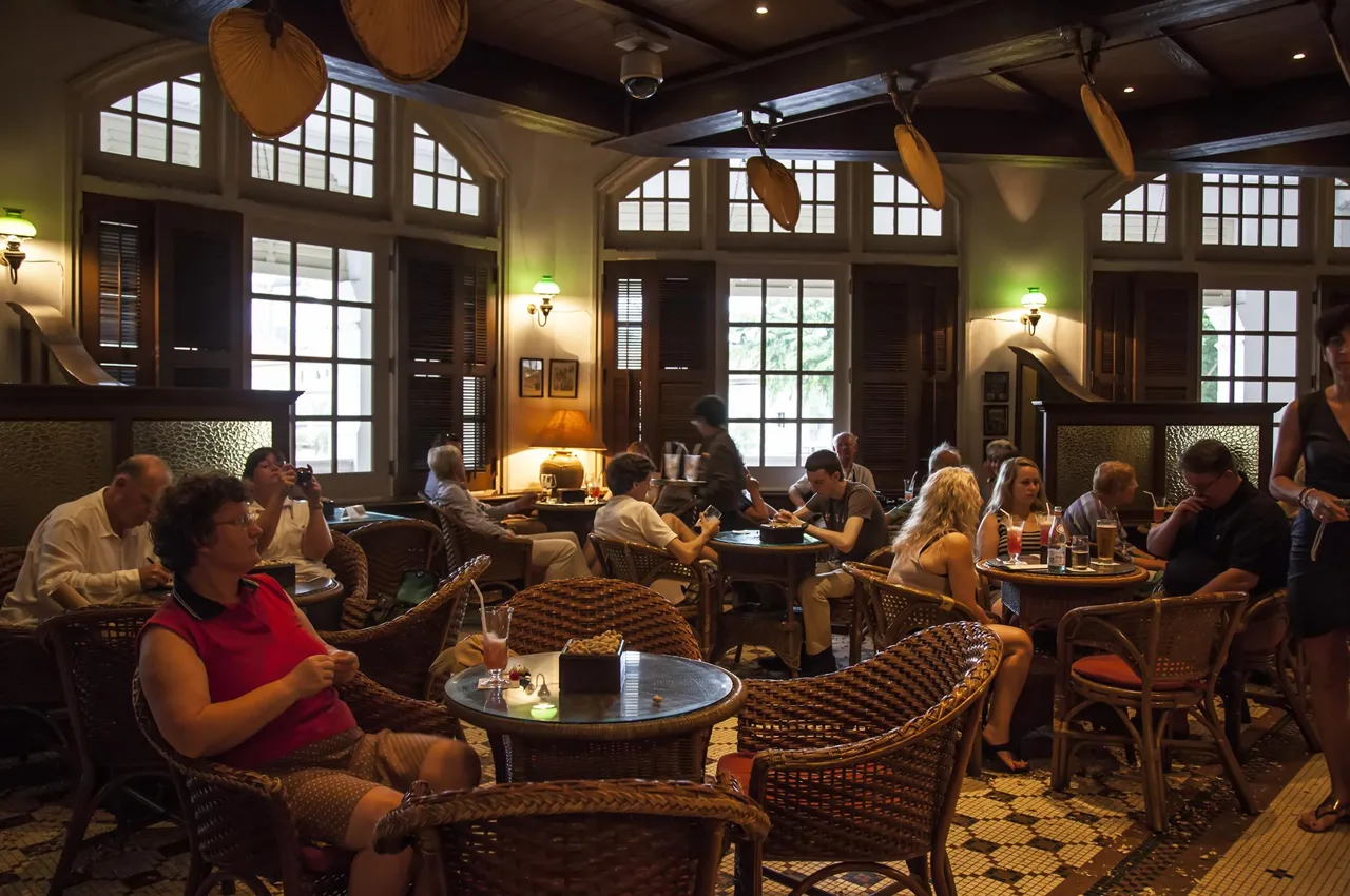 Inside the Long Bar: woven fans hanging from the beams, rattan armchairs around small round tables, a patterned tile floor, and seated guests.