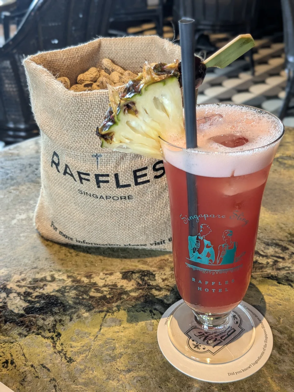 A Singapore Sling in a tall printed glass with a pineapple wedge and a cherry, beside a hessian sack of peanuts marked RAFFLES SINGAPORE.