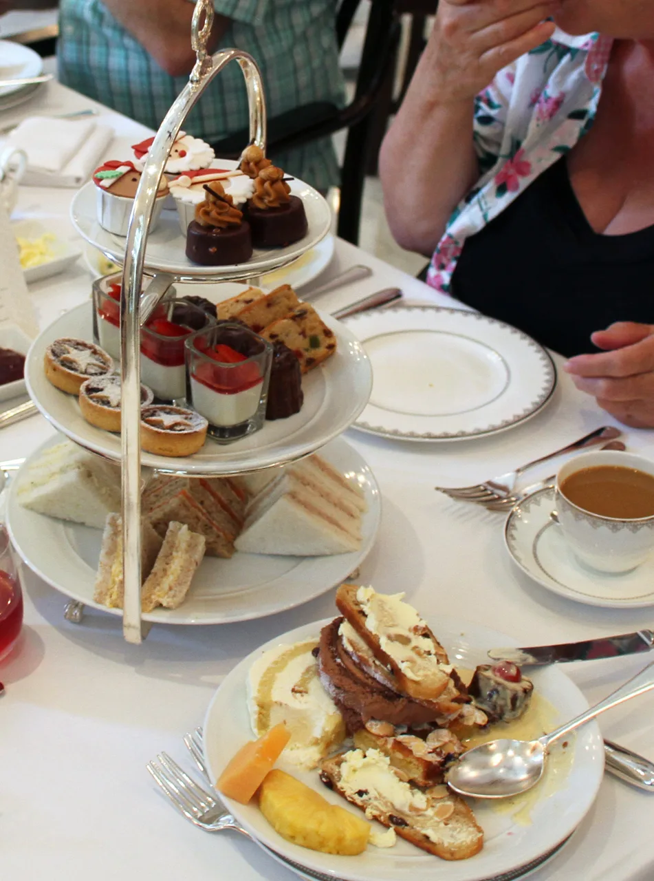 A three-tier silver stand on white linen holding finger sandwiches, scones and small pastries, with a teacup beside it.