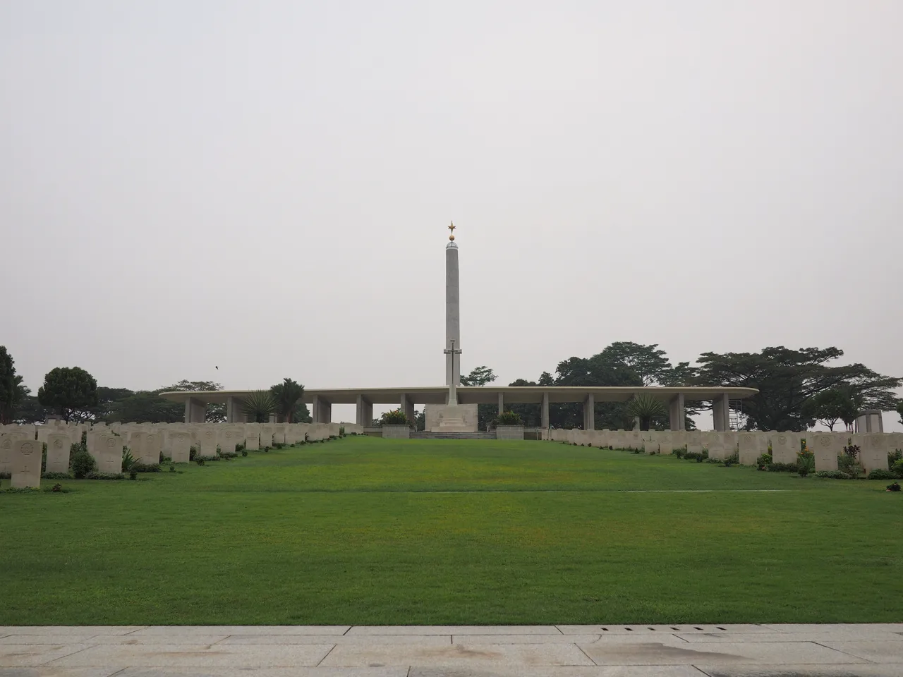 White headstones in rows on both sides of a lawn at Kranji War Cemetery, with a pale pylon and a long low roofed memorial behind them
