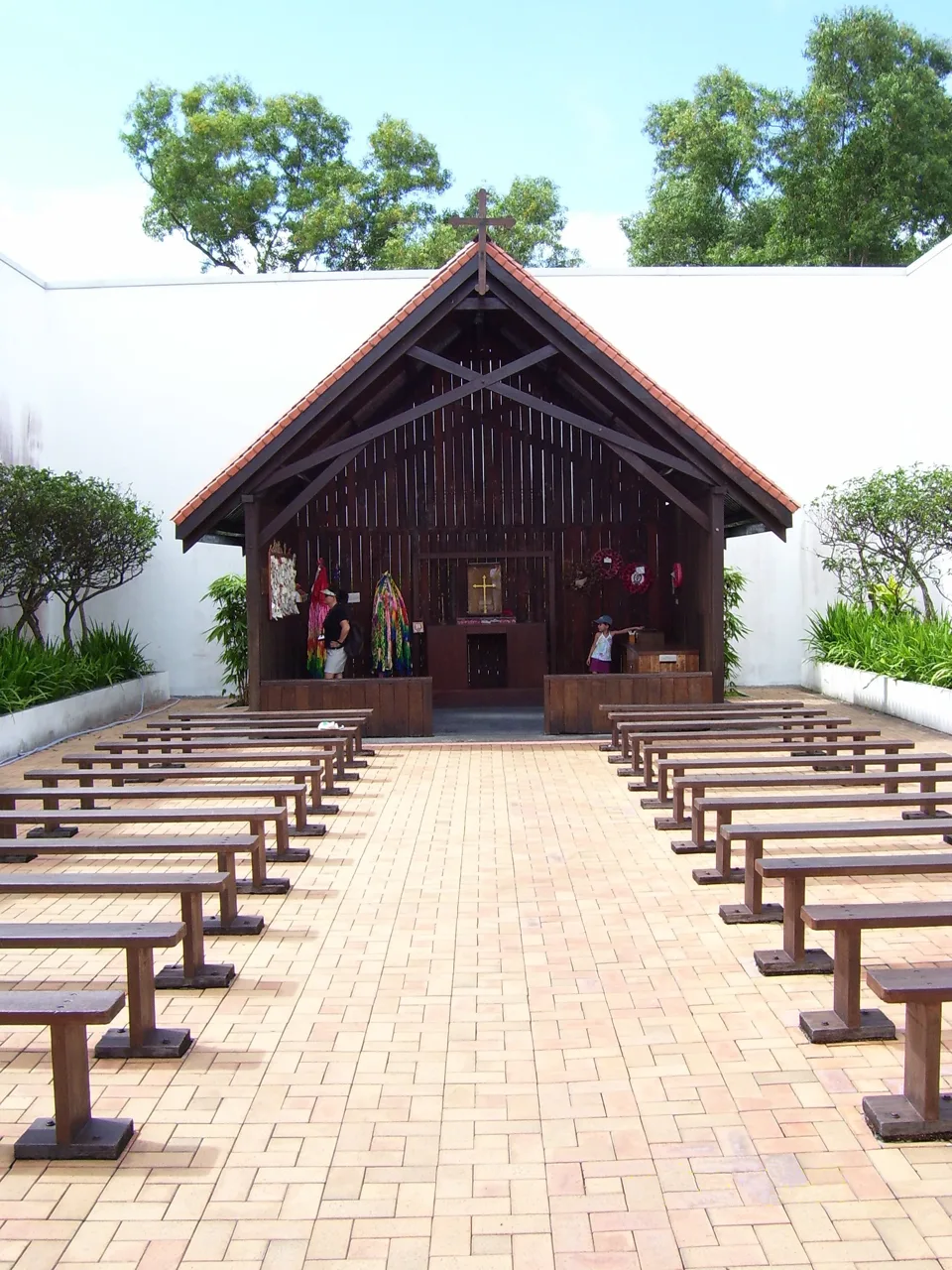 An open-fronted wooden chapel with a cross on its roof, with rows of wooden benches set out in front of it