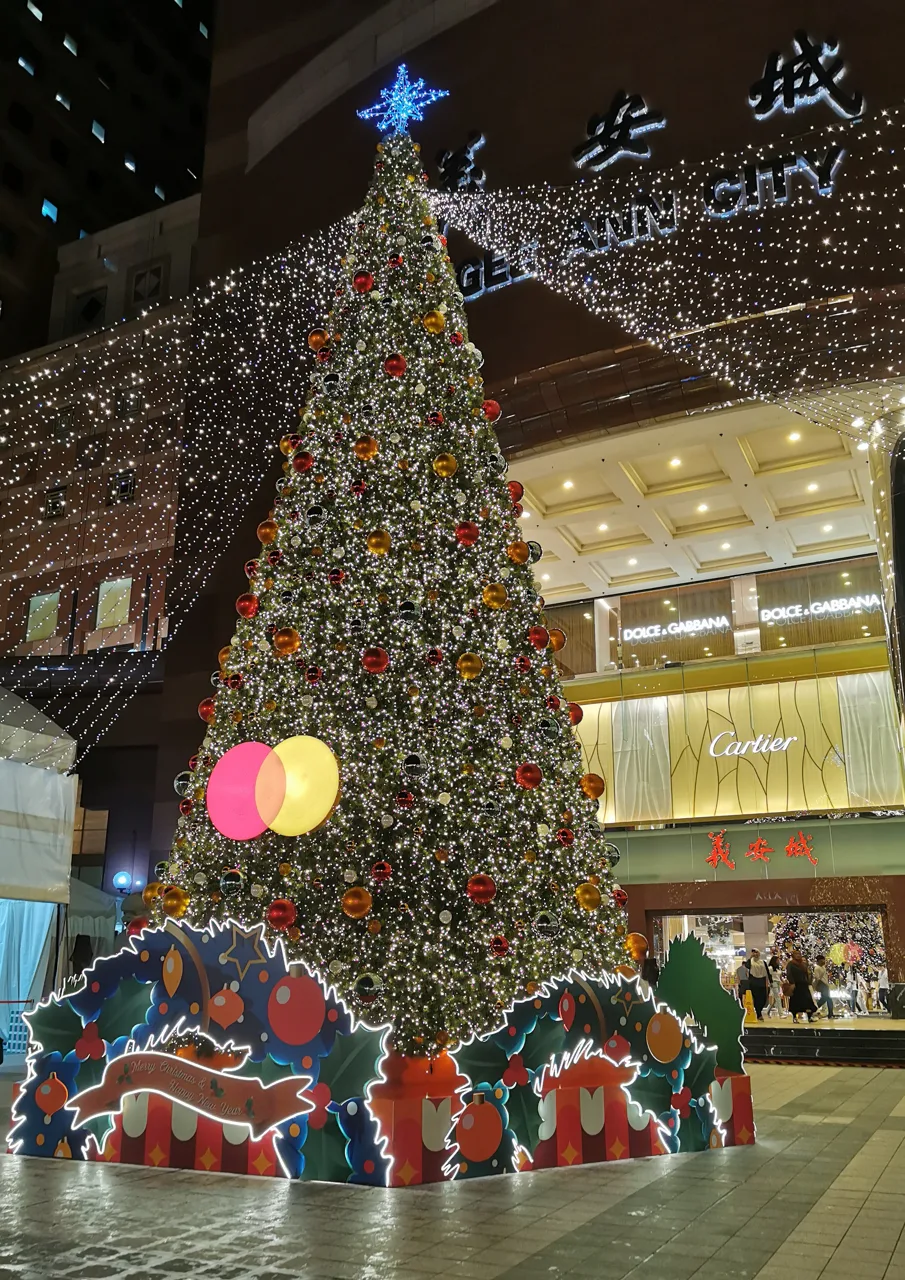 A tall Christmas tree hung with baubles and lights in front of Ngee Ann City at night, with curtains of lights on the building.
